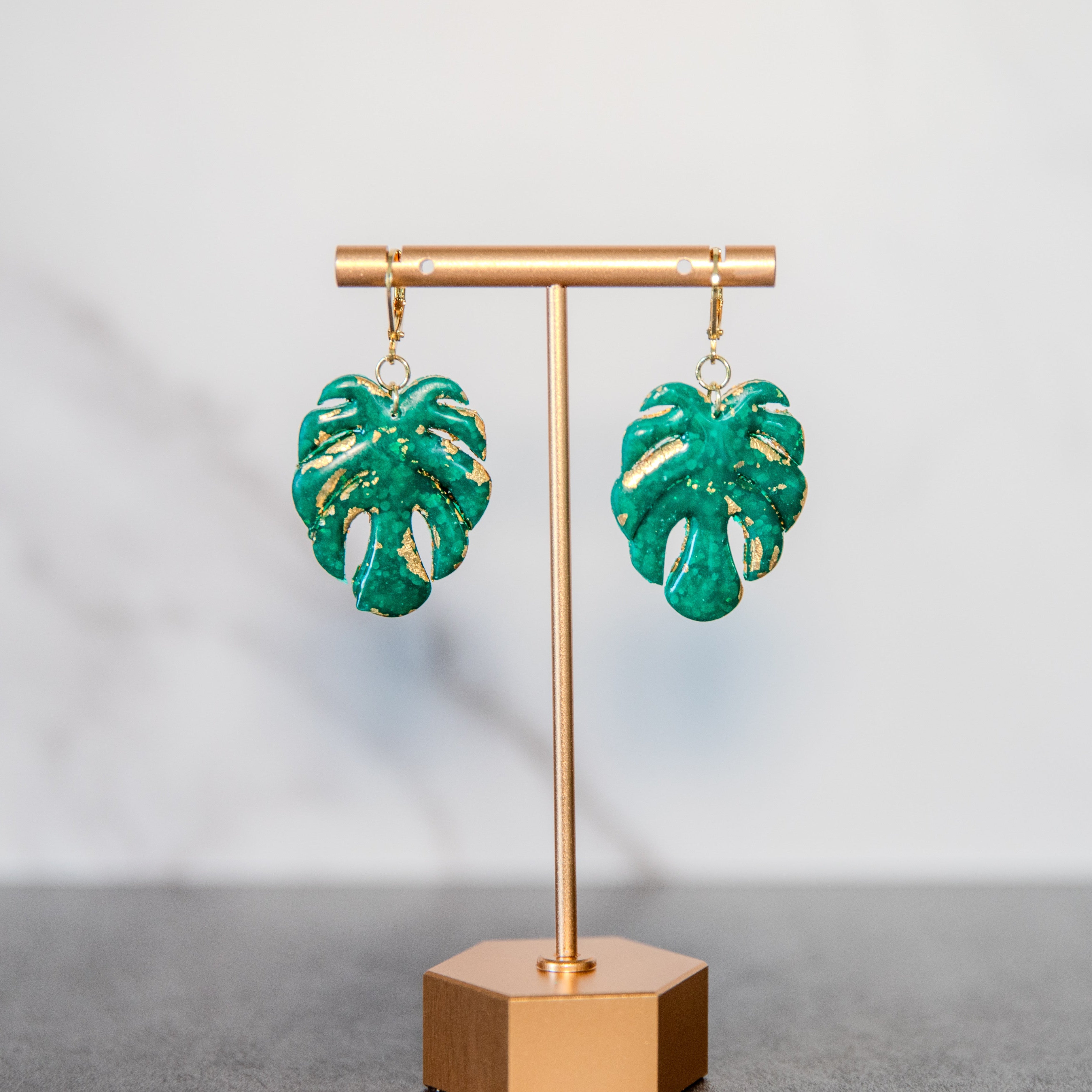 Green leaf-shaped earrings on a gold stand against a gray background