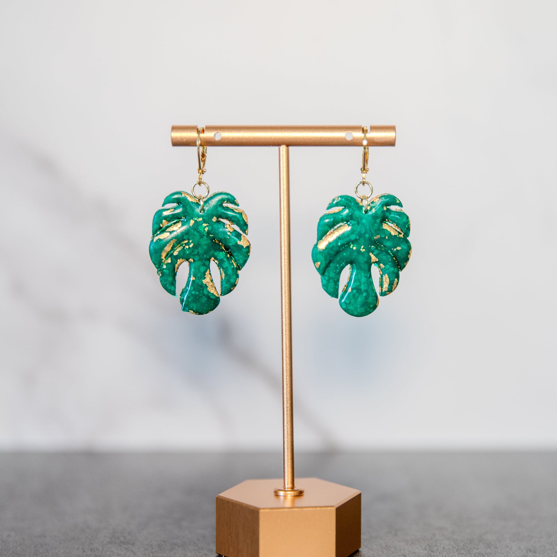 Green leaf-shaped earrings on a gold stand against a gray background