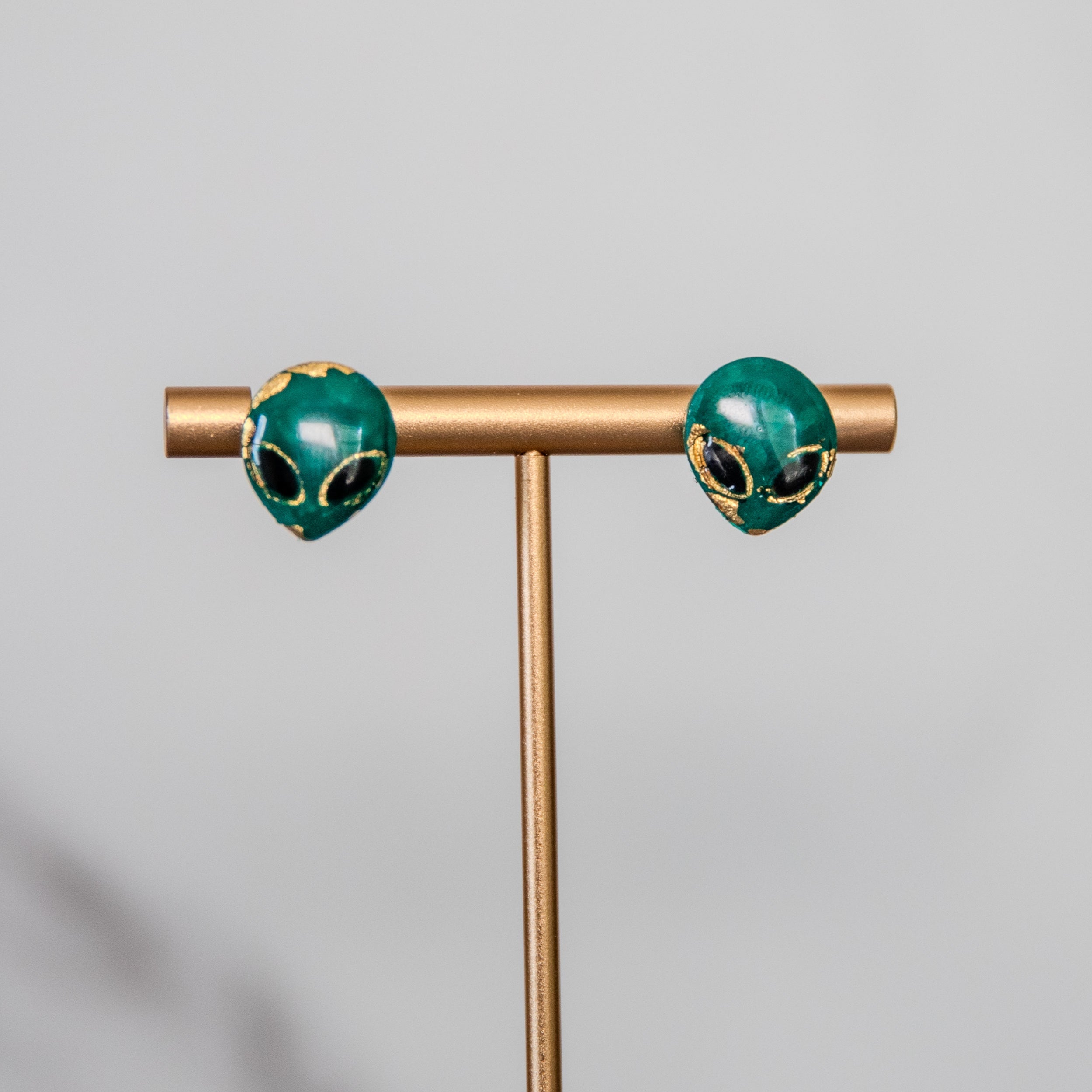 Pair of green alien earrings on a gold T-shaped stand against a gray background