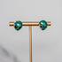 Pair of green alien earrings on a gold T-shaped stand against a gray background