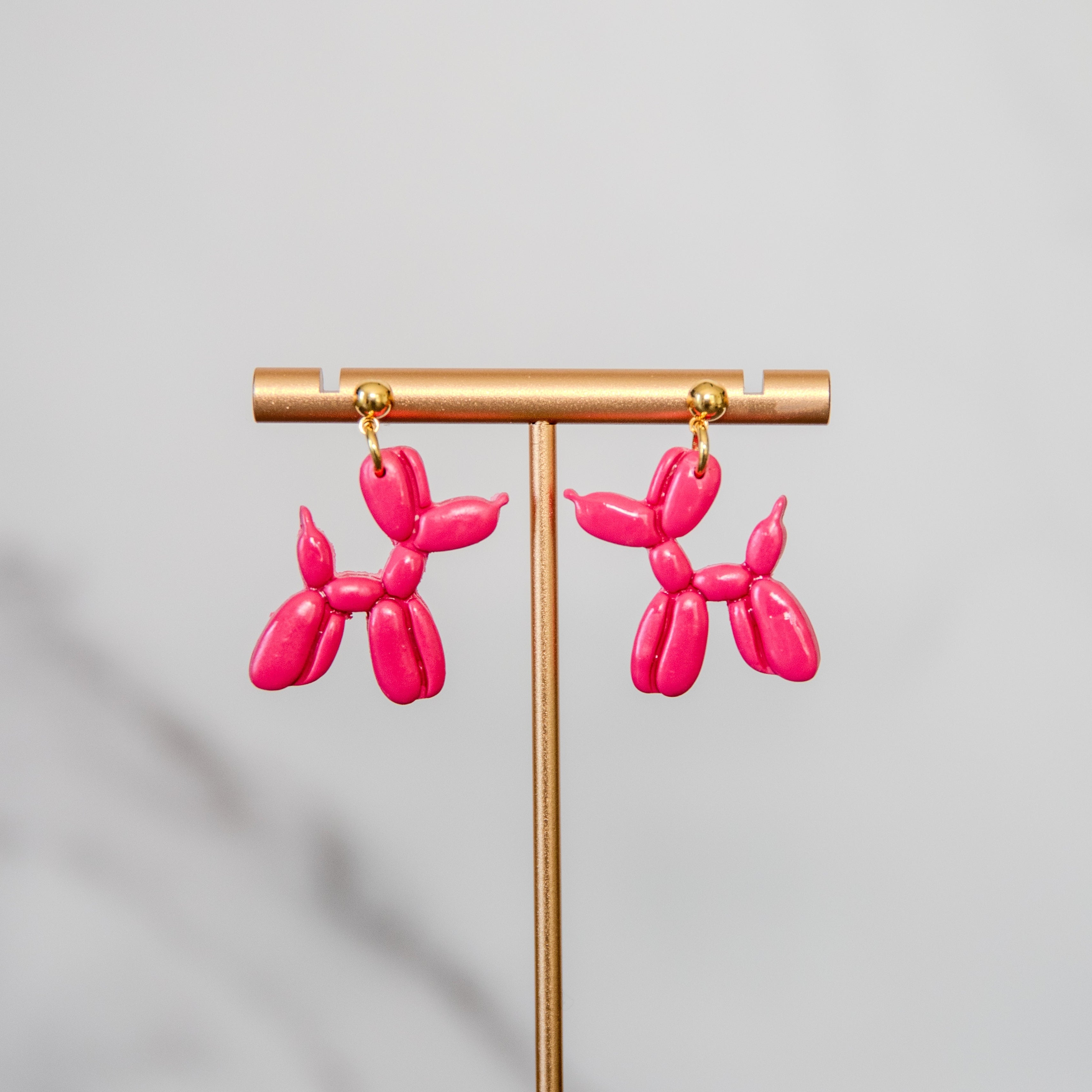 Pink balloon dog-shaped earrings on a gold stand against a gray background