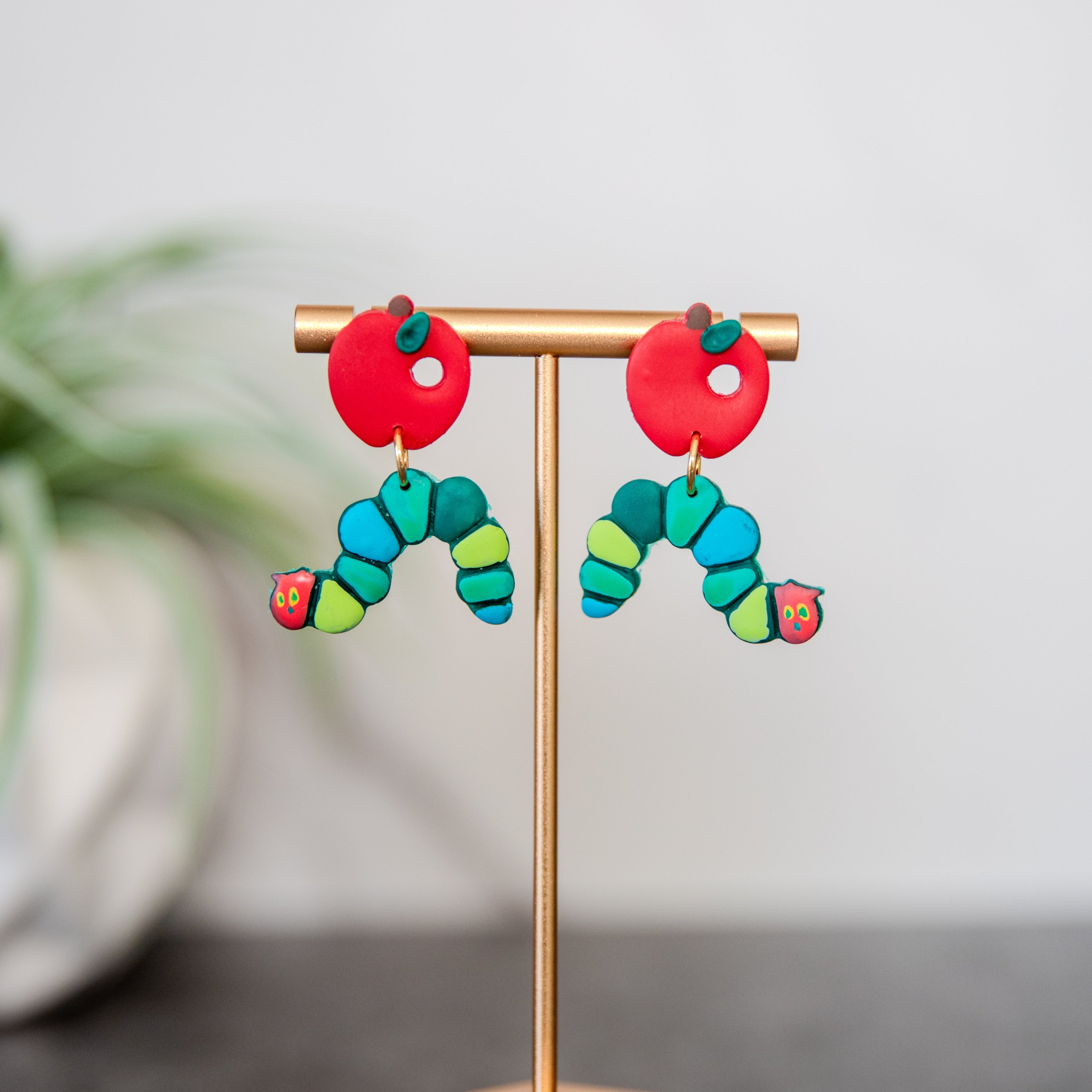 Colorful caterpillar-shaped earrings on a stand with a plant in the background