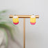 Pair of pencil earrings on a gold stand with a blurred plant in the background