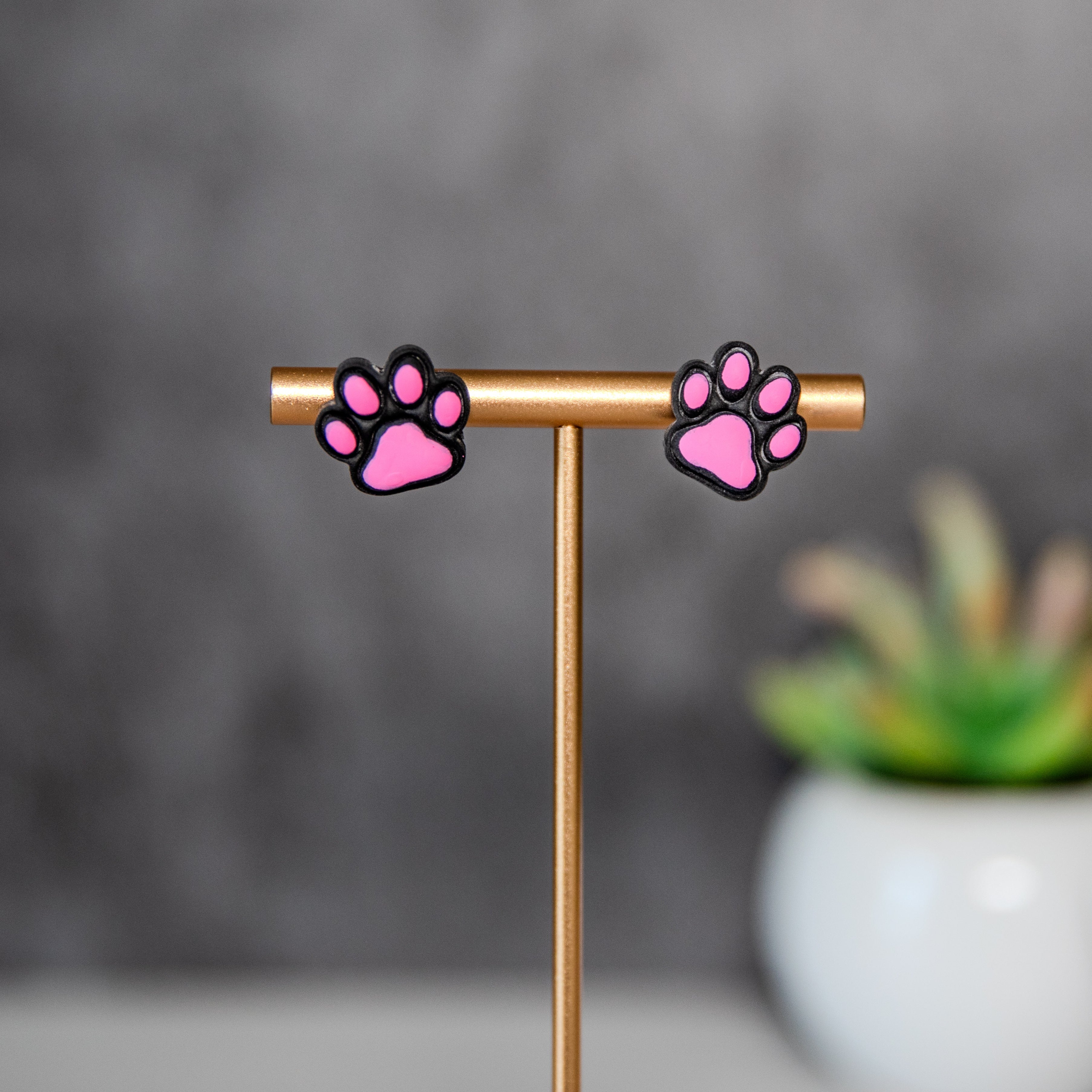 Pink paw print earrings on a gold stand with a blurred plant in the background