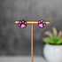 Pink paw print earrings on a gold stand with a blurred plant in the background