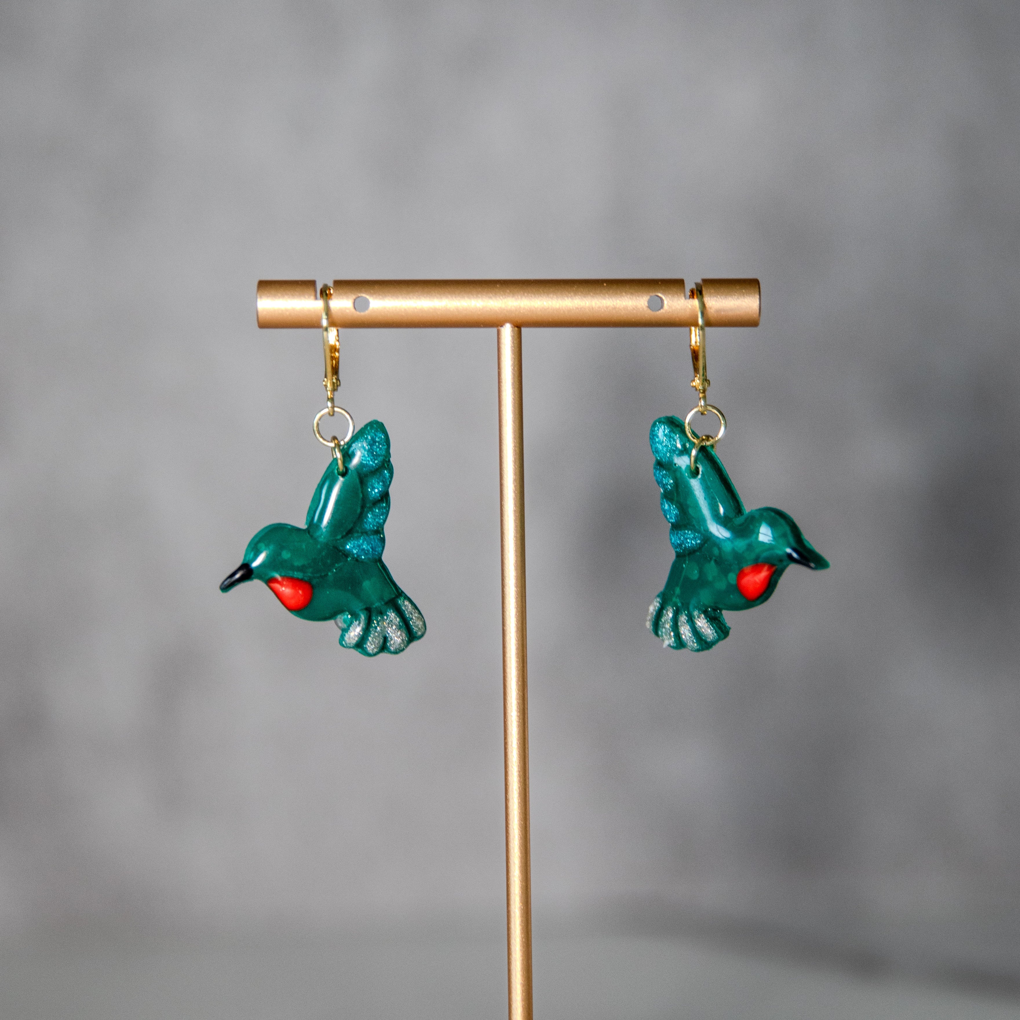 Pair of green bird-shaped earrings on a gold stand against a gray background