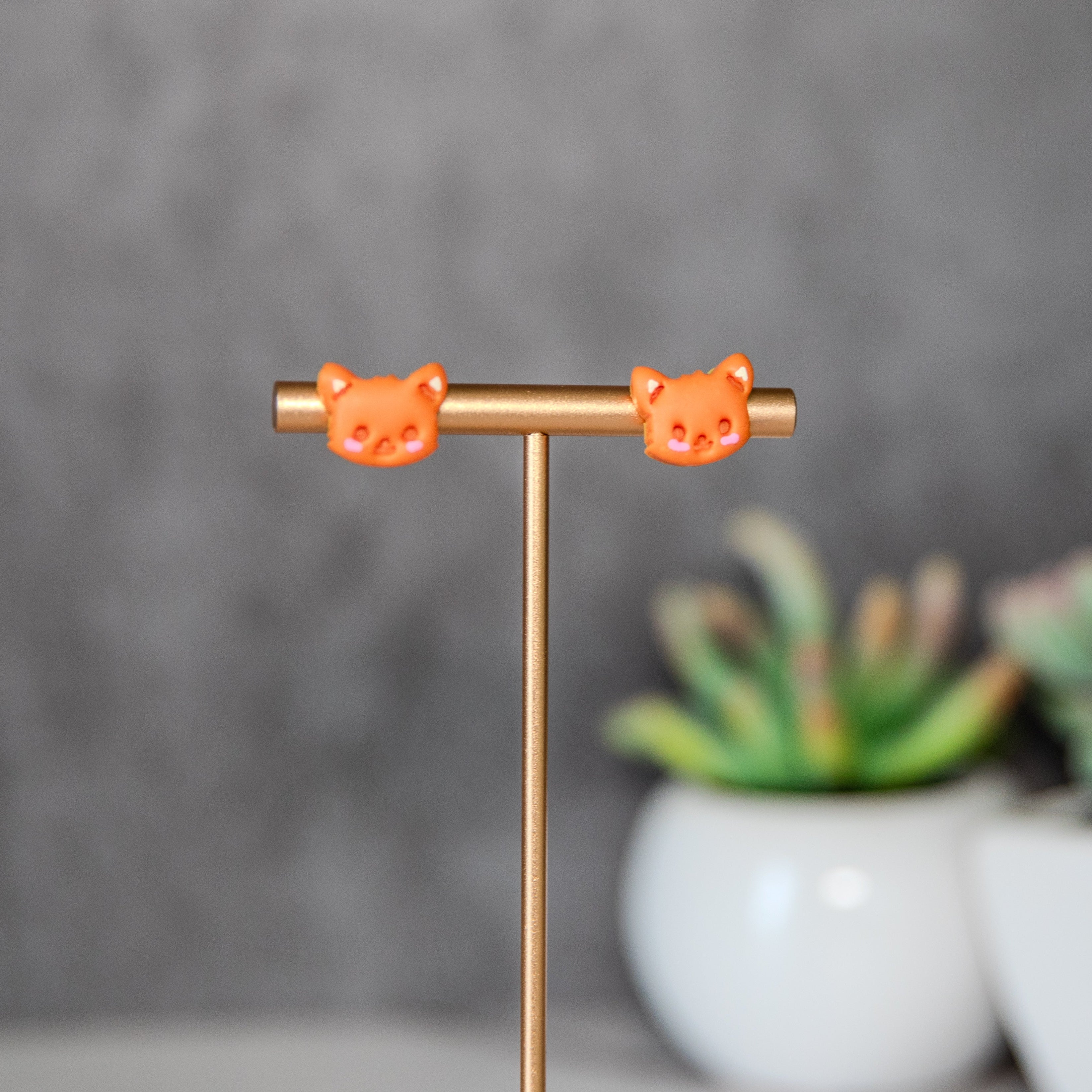 Orange cat-shaped earrings on a gold T-shaped stand with a blurred background