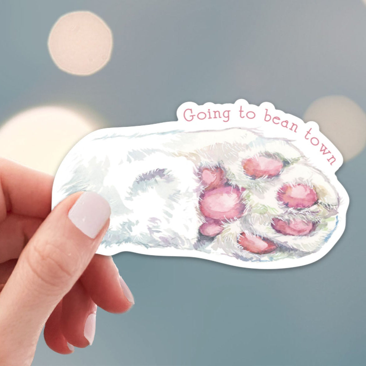 Hand holding a sticker with cat paw print design and text 'Going to bean town' against a blurred background