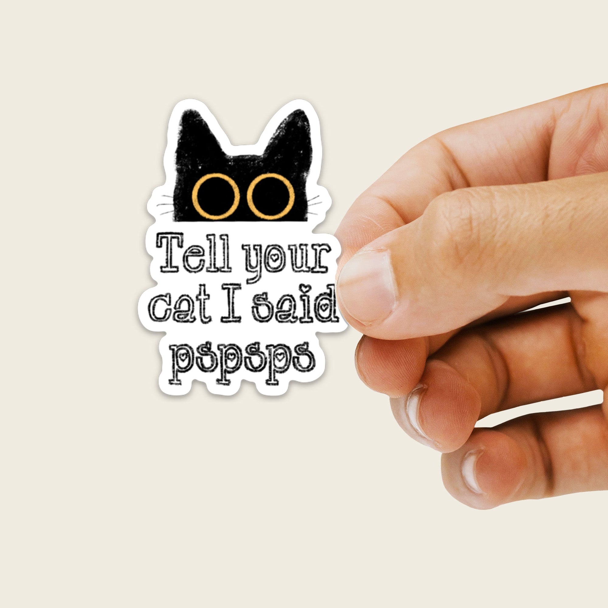 Hand holding a sticker with a cat face and text on a beige background