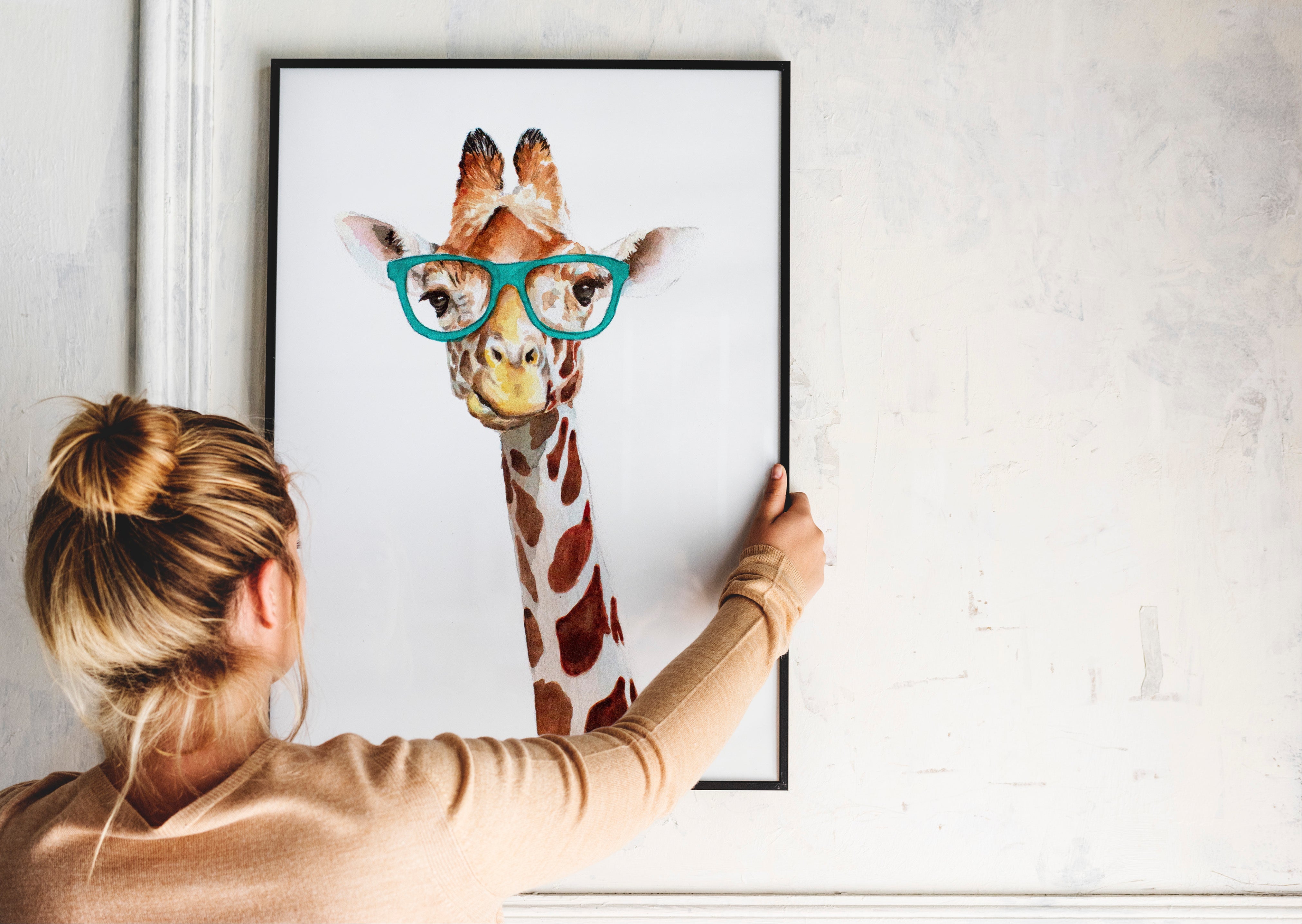 Person hanging a framed picture of a giraffe wearing glasses on a wall.