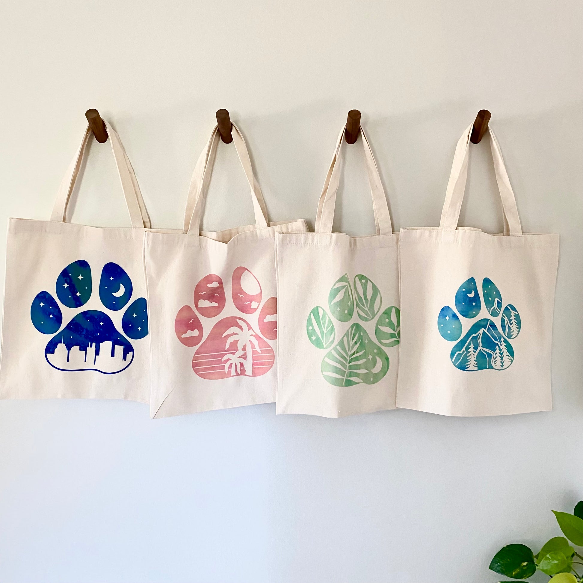 Four tote bags with paw print designs hanging on a wall.