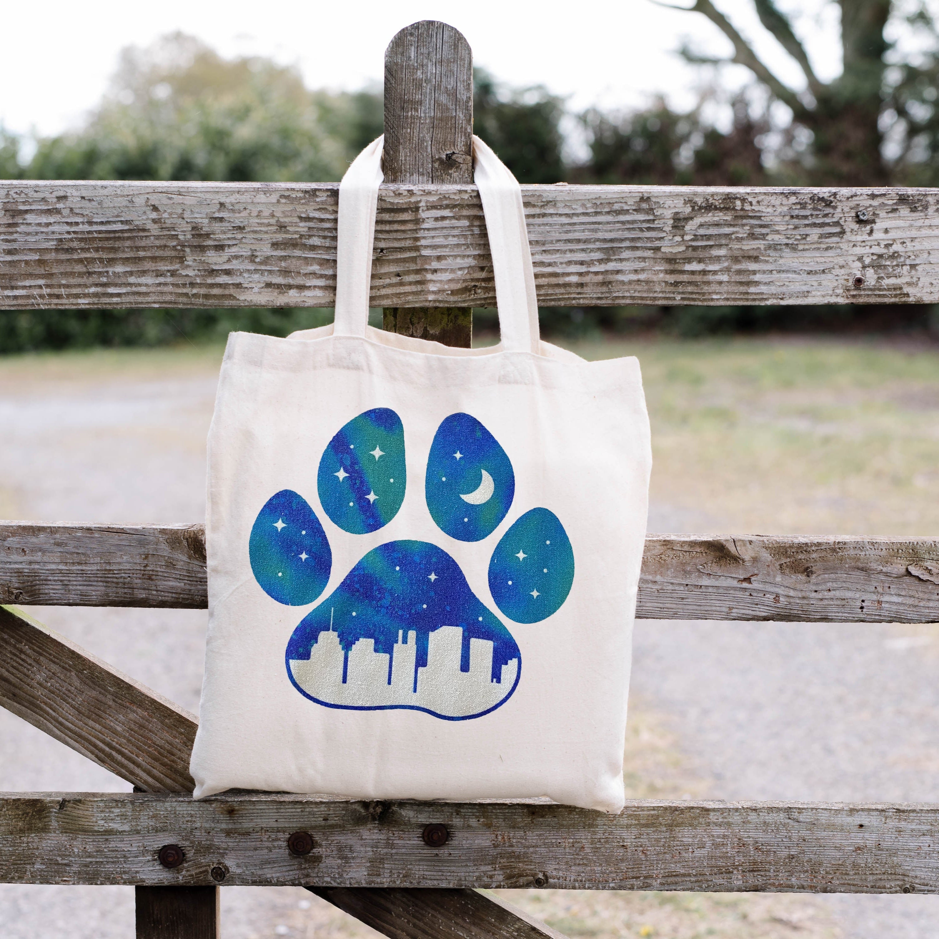 Tote bag with a blue paw print design hanging on a wooden fence outdoors.