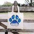 Tote bag with a blue paw print design hanging on a wooden fence outdoors.