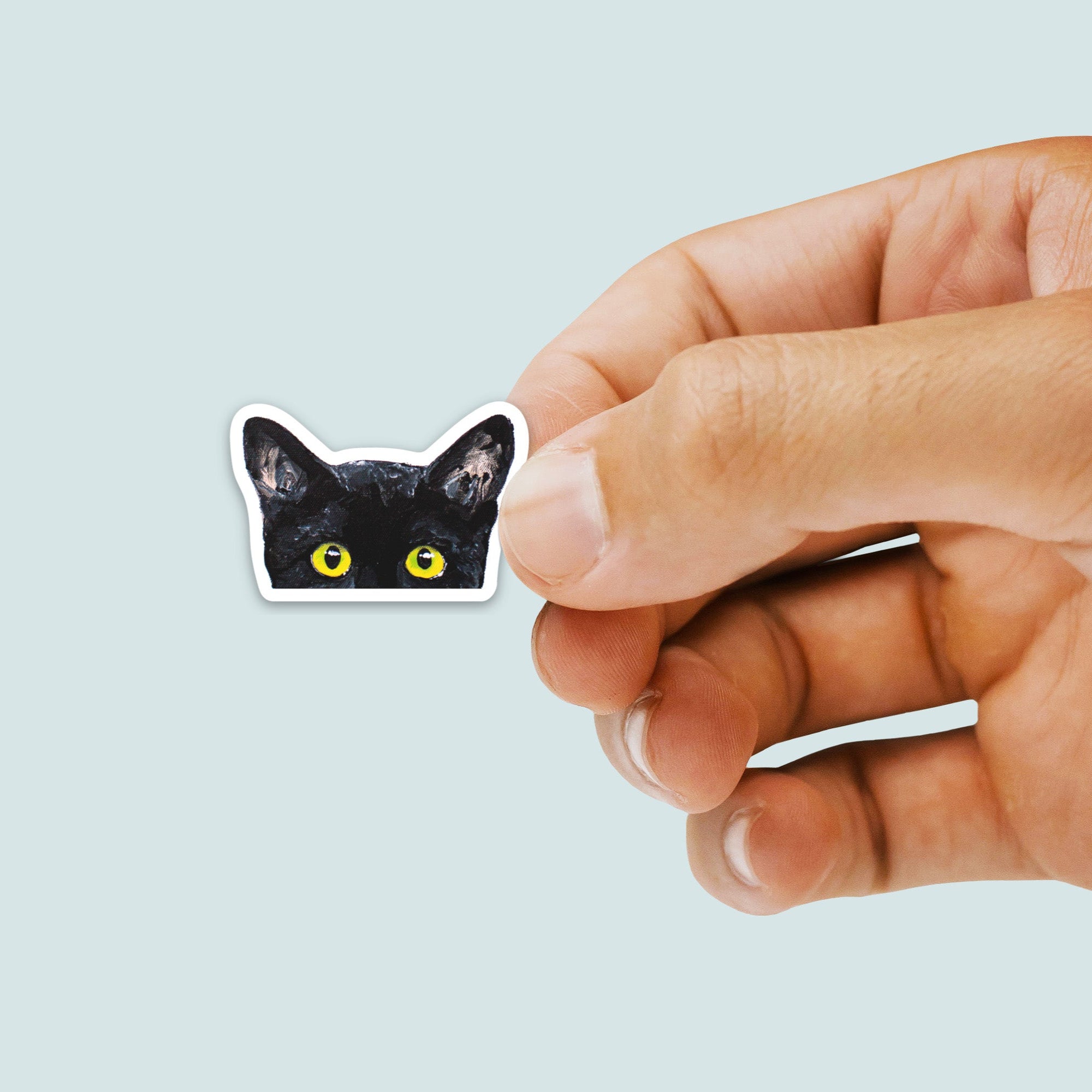 Hand holding a black cat sticker with yellow eyes on a light gray background