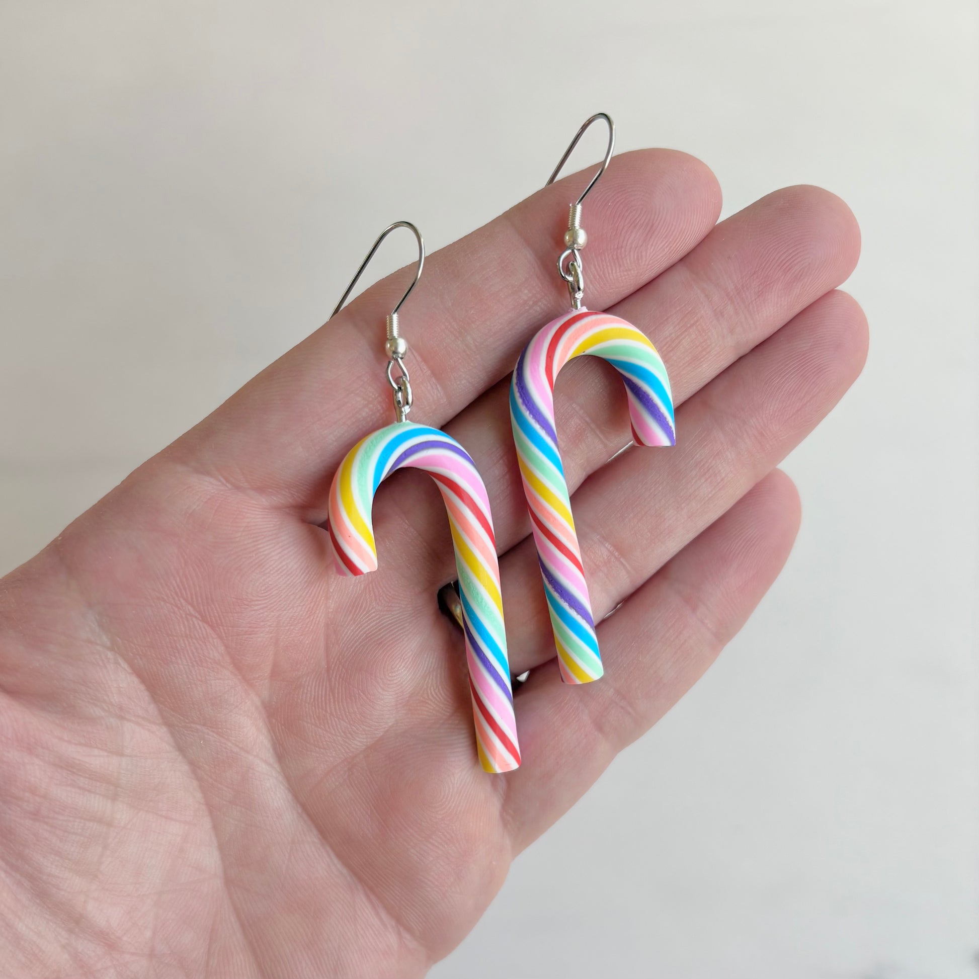 Rainbow candy cane-shaped earrings held in a hand against a plain background