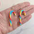 Rainbow candy cane-shaped earrings held in a hand against a neutral background