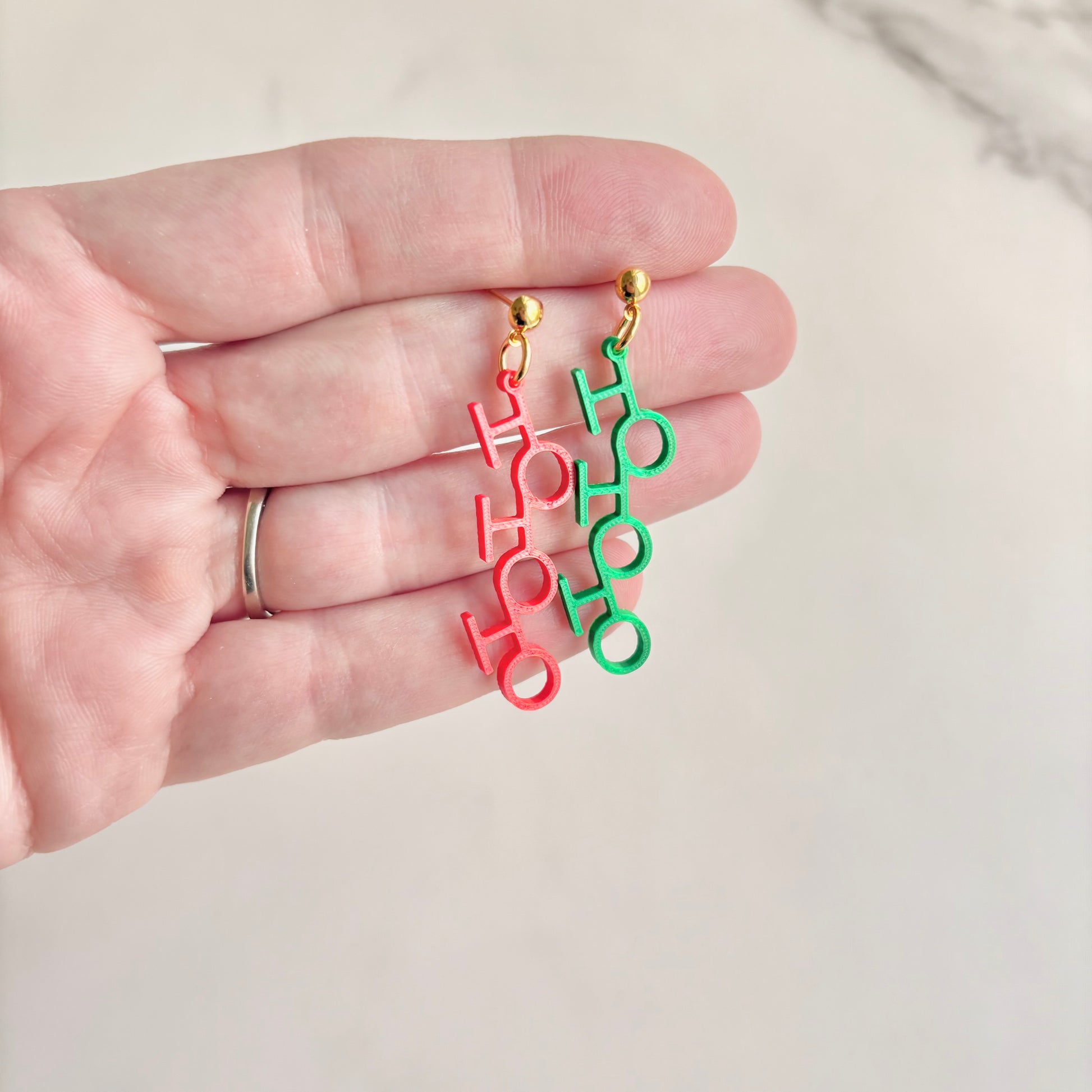 Colorful 'Ho Ho Ho' earrings held in a hand against a neutral background