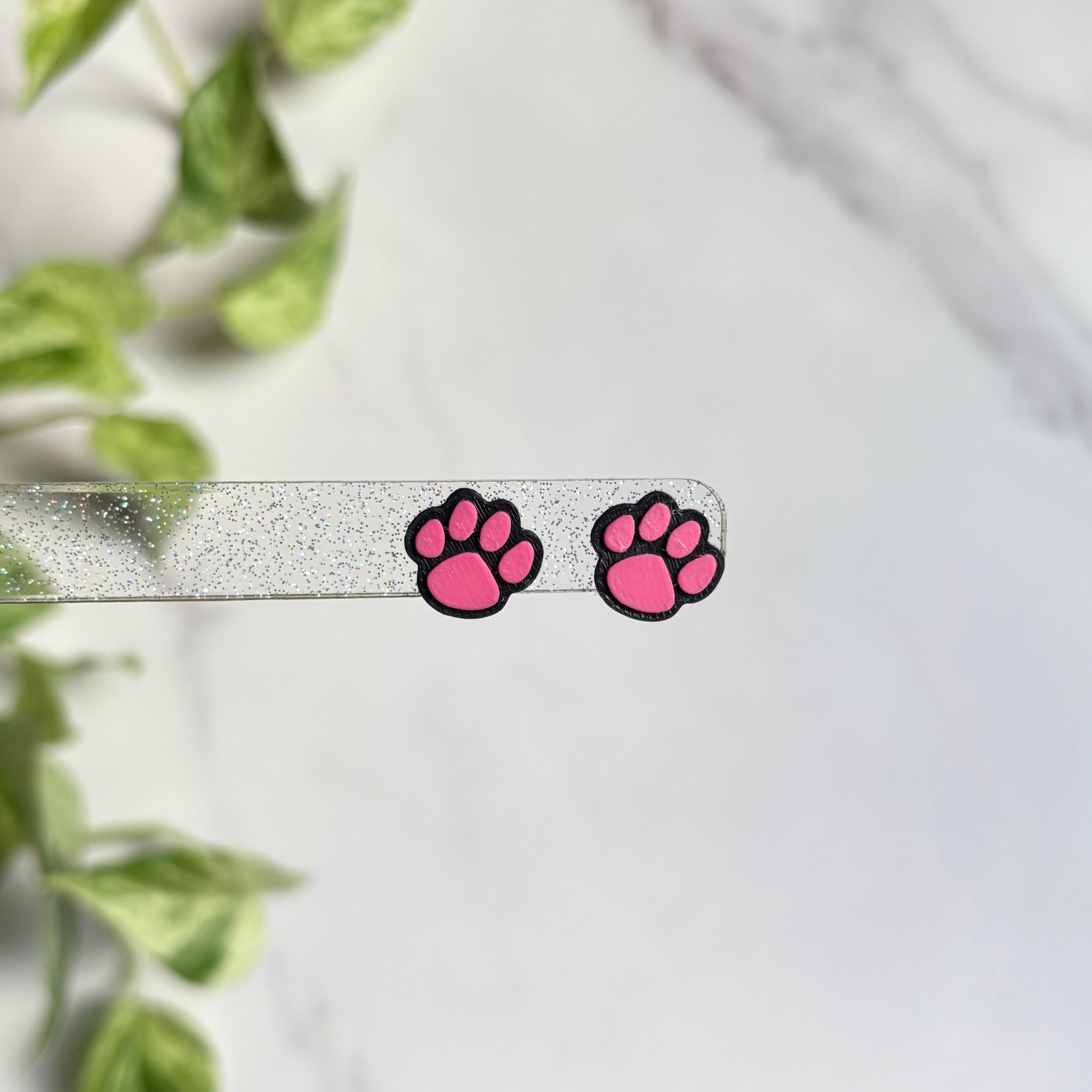 Pink paw print earrings on a white background with green leaves in the corner