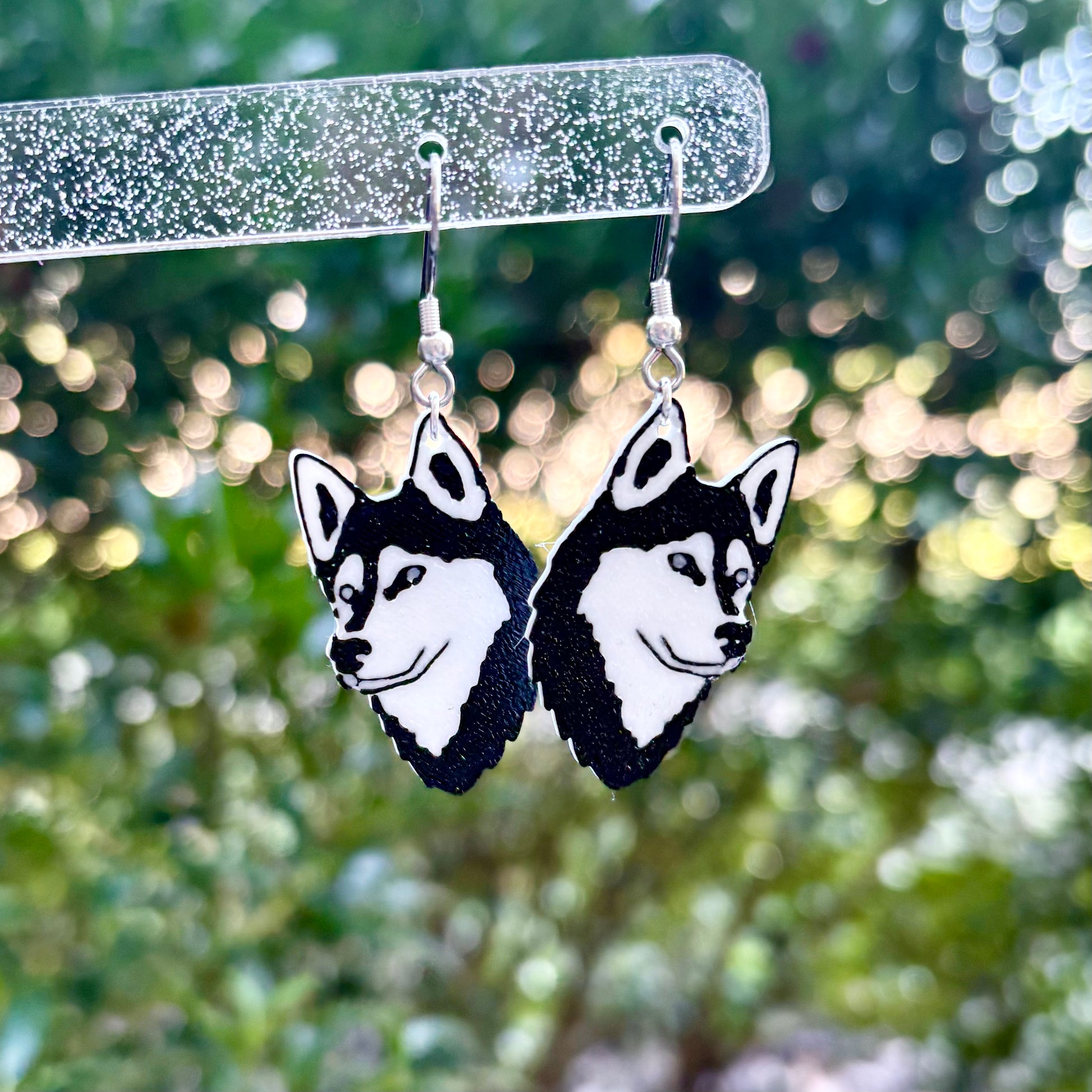 Husky dog face earrings hanging on a clear plastic stand with a blurred green background.