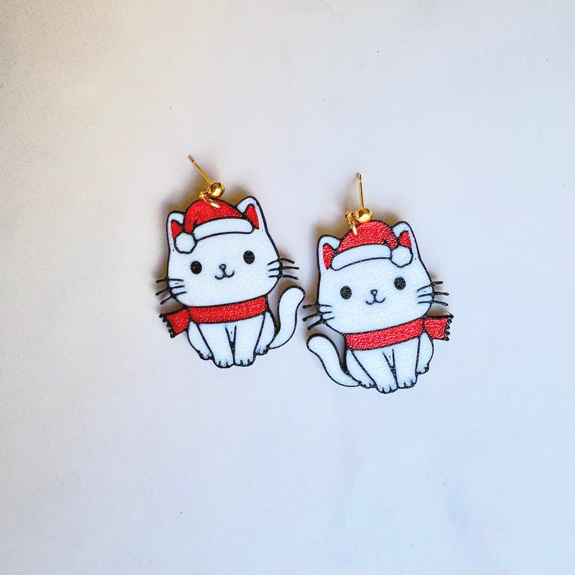 Pair of cat-shaped earrings with red scarves and hats on a white background