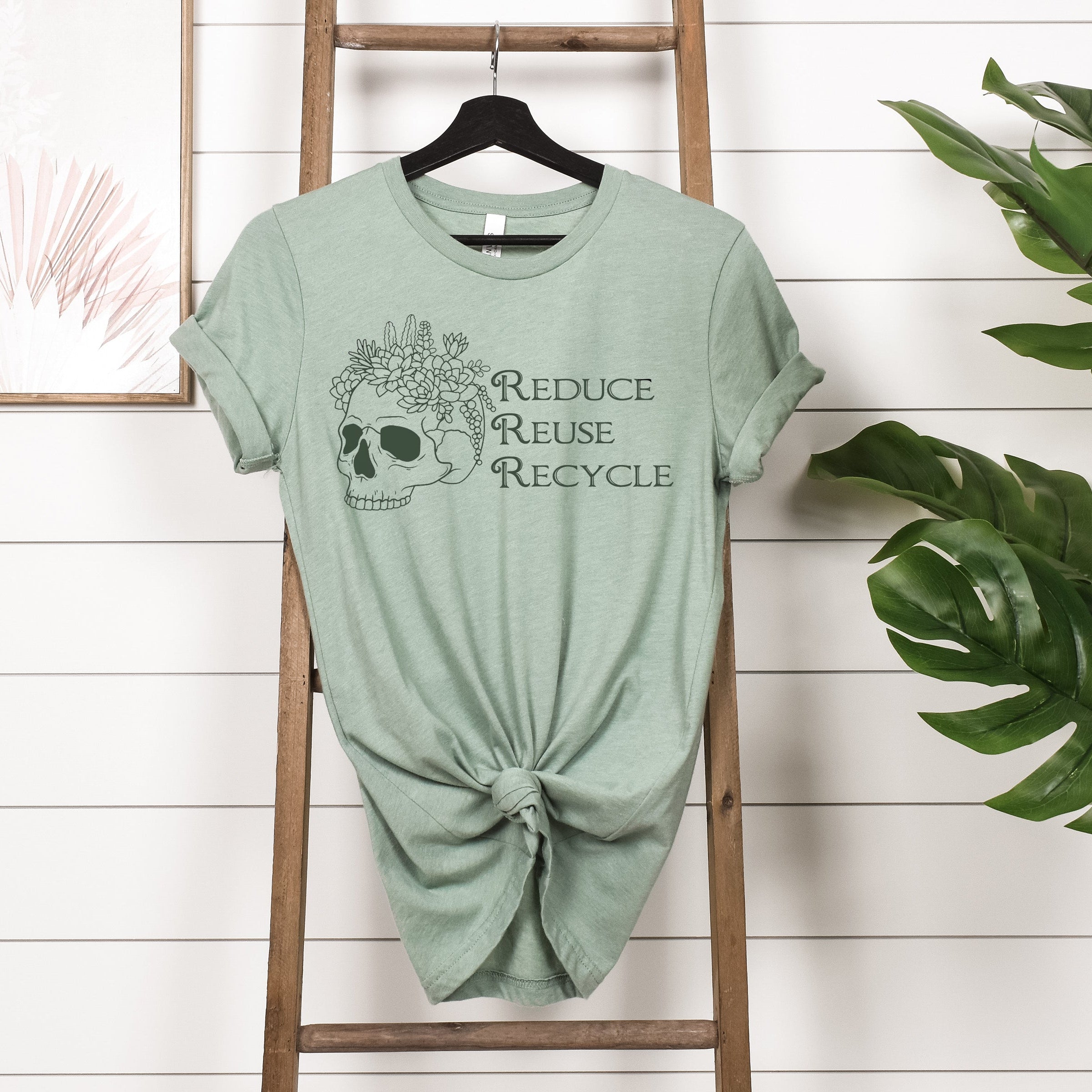 Green t-shirt with skull design and text on a wooden ladder against a white wall with plants.