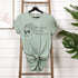 Green t-shirt with skull design and text on a wooden ladder against a white wall with plants.